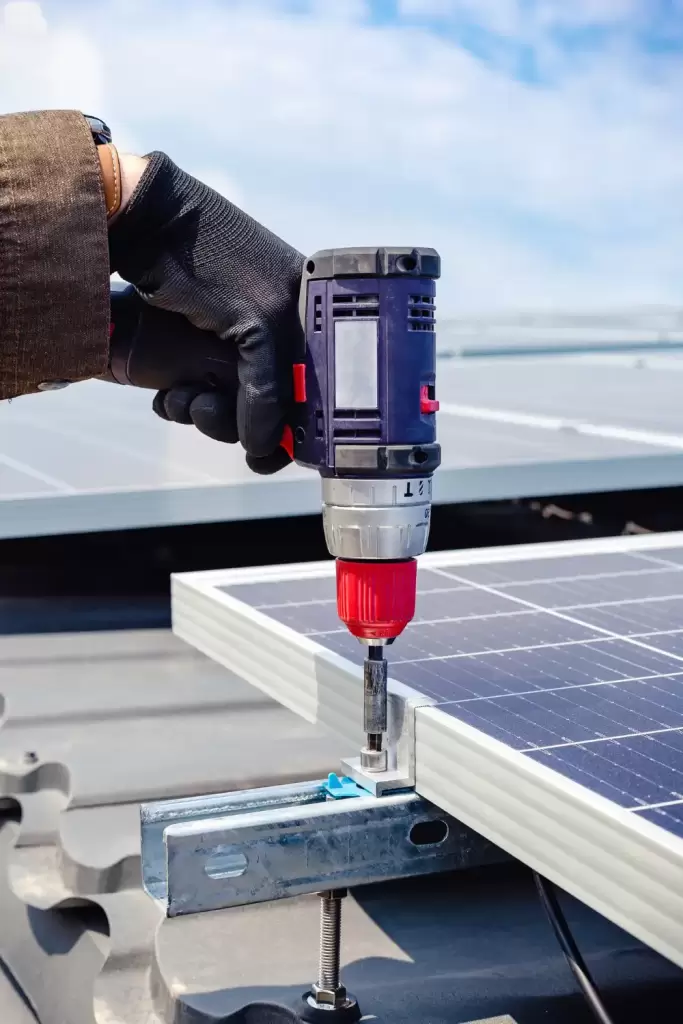 Bir teknisyen, güneş santralinin montajını yaparken. Teknisyen, panelleri monte ederken güvenlik donanımı kullanıyor ve arkasında berrak mavi gökyüzü ile güneş panelleri görünüyor.