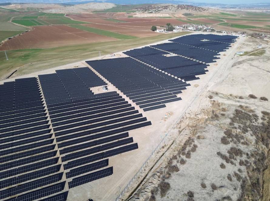 Güneş enerjisi sistemleri kurulmuş bir tarlada, panellerin ve çevresel yeşilliklerin gösterildiği fotoğraf.